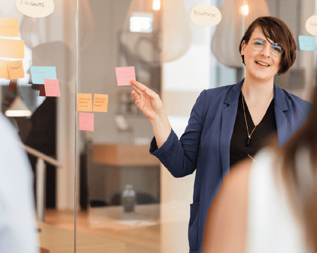 Yvonne-Homann-erarbeitet-mit-ihren-Kunden-eine-Markenstrategie-sie-steht-an-einer-Glaswand-und-klebt-ein-Post-it-mit-Ergebnissen-des-Markenworkshops-an-die-Wand