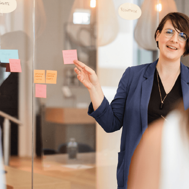 Yvonne-Homann-erarbeitet-mit-ihren-Kunden-eine-Markenstrategie-sie-steht-an-einer-Glaswand-und-klebt-ein-Post-it-mit-Ergebnissen-des-Markenworkshops-an-die-Wand